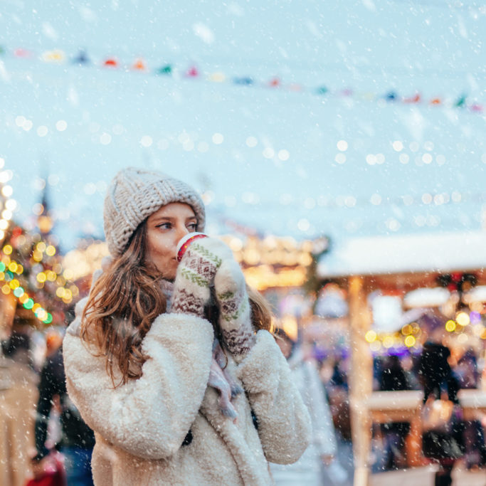 Girl drinking hot coffee while walking in Christmas market decorated with holiday lights in the evening. Feeling happy in big city. Spending winter vacations in Red square, Moscow, Russia.